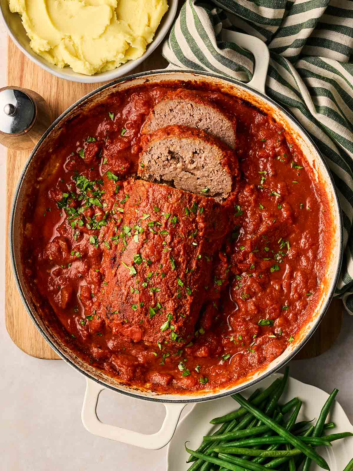 A large pan filled with a meatloaf in a rich tomatoey sauce, on a table with mashed potato and green beans to the side, ready to serve.