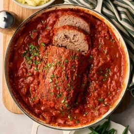 A large of of tomato sauce with a meatloaf in the middle, with a few slices and parsley over the top.