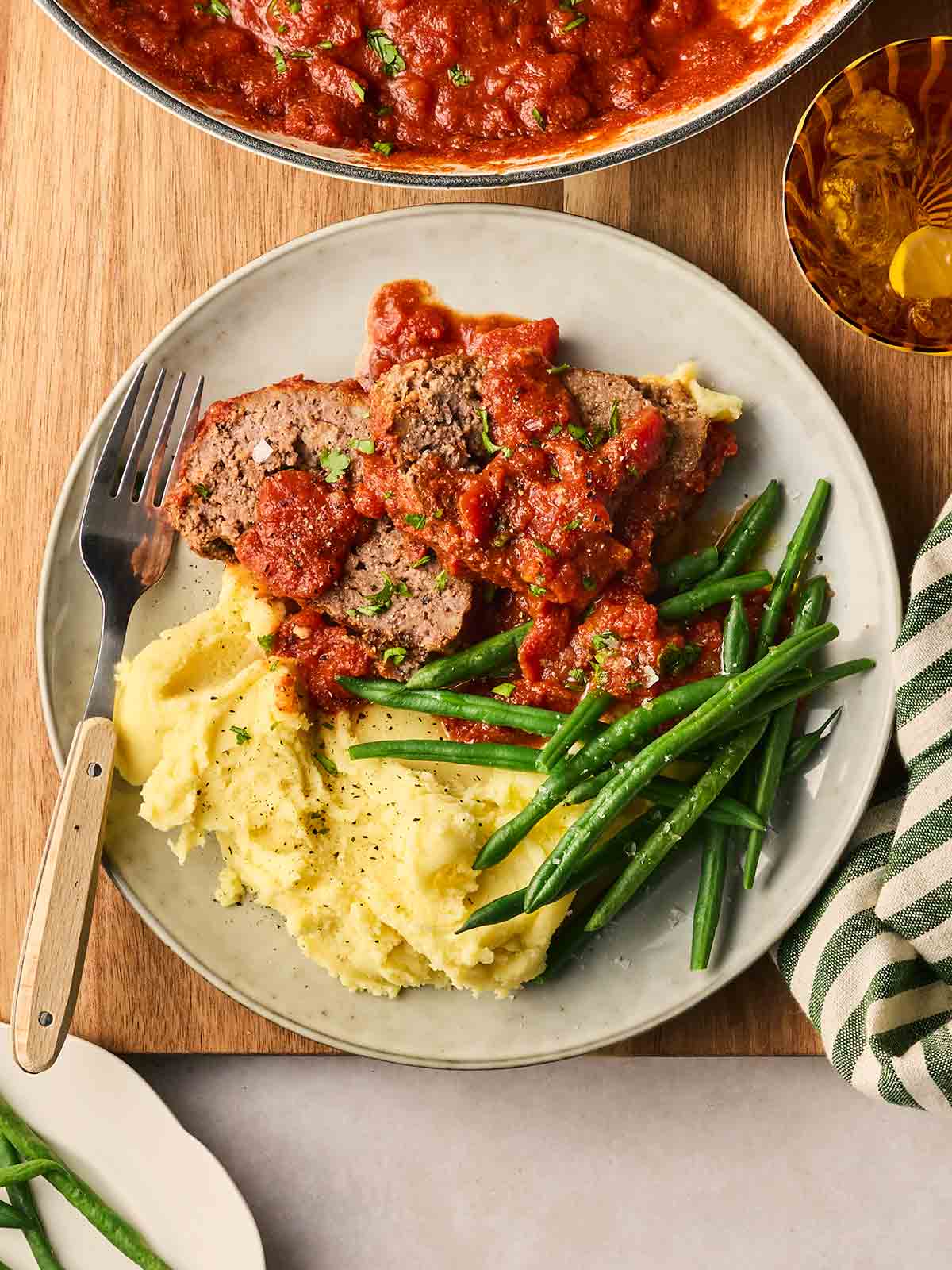 A plate on a table with slices of meatloaf in a tomato sauce, green beens and mashed potatoes, with a fork ready to serve.