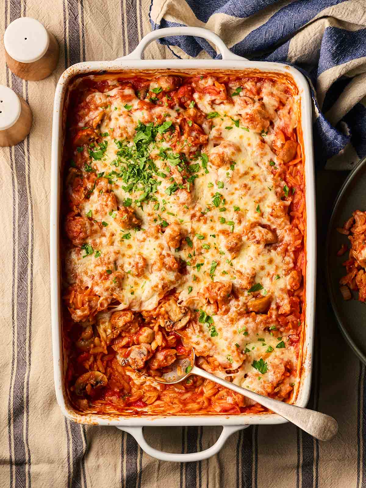 An oven dish filled with cooked Sausage Orzo Bake on a table, ready to serve with a spoon.