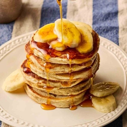 A stack of pancakes on a plate with banana slices and maple syrup being poured from a jug.