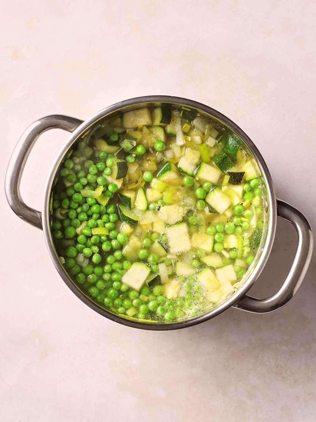 A metal pan with cooking vetetables, including peas and courgettes cooking in stock.