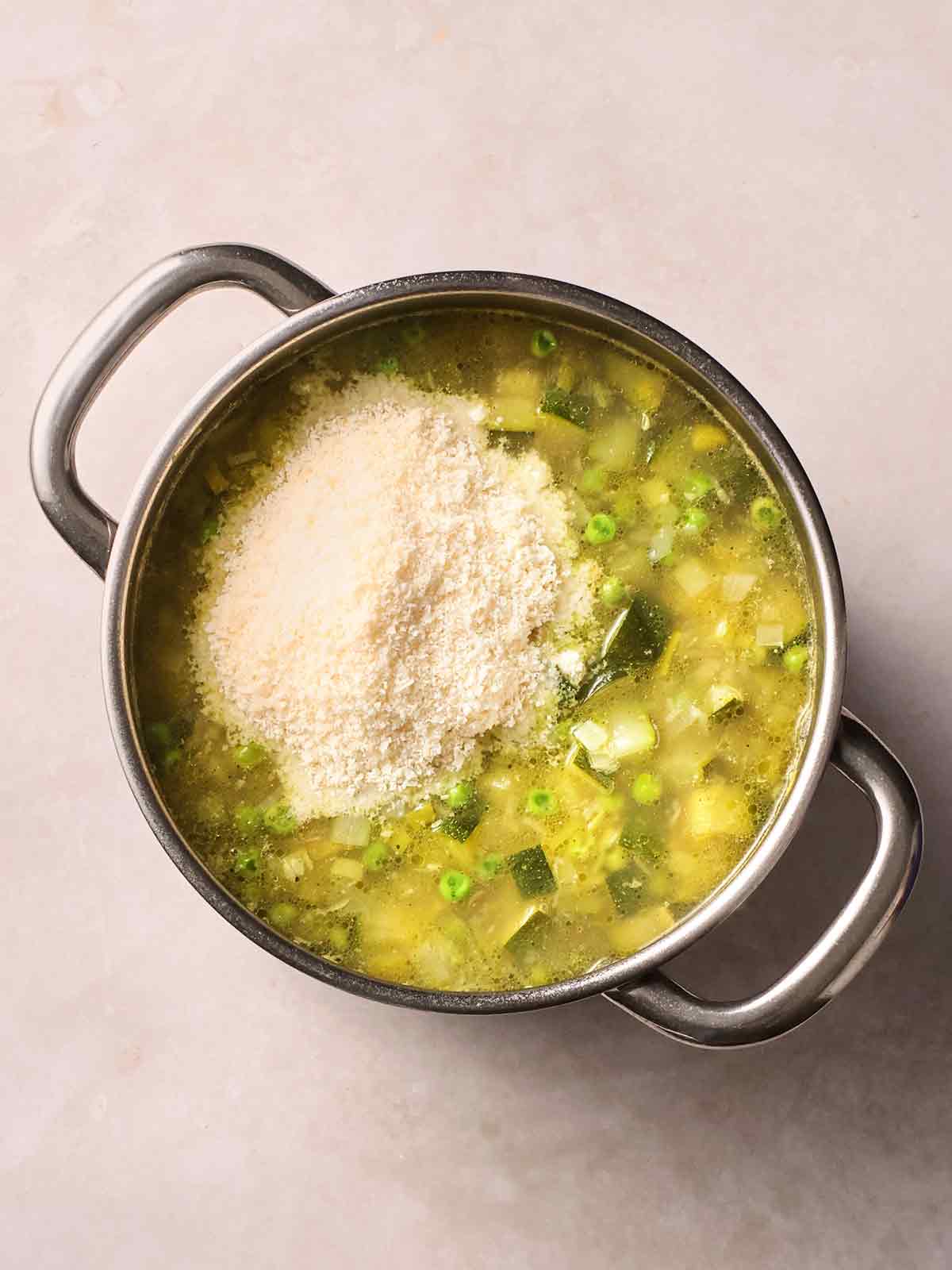 A metal large pan with vegetables cooking in a soup, with a pile of parmesan cheese on top, ready to mix in.