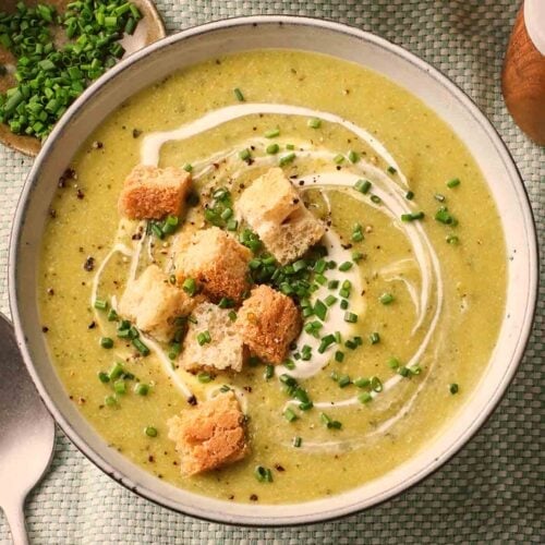 A green soup on a table, with croutons and chives on top, ready to eat with a spoon to the side.