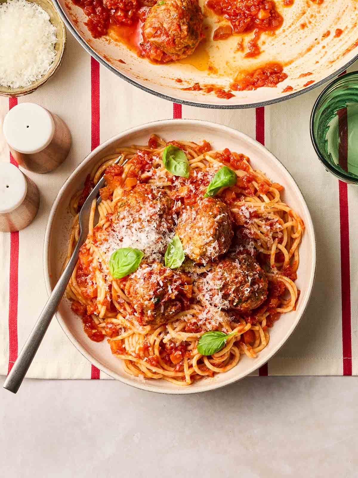 A bowl filled with classic homemade meatballs with spaghetti and a fork, with a large pan to the side, on a set table for dinner.