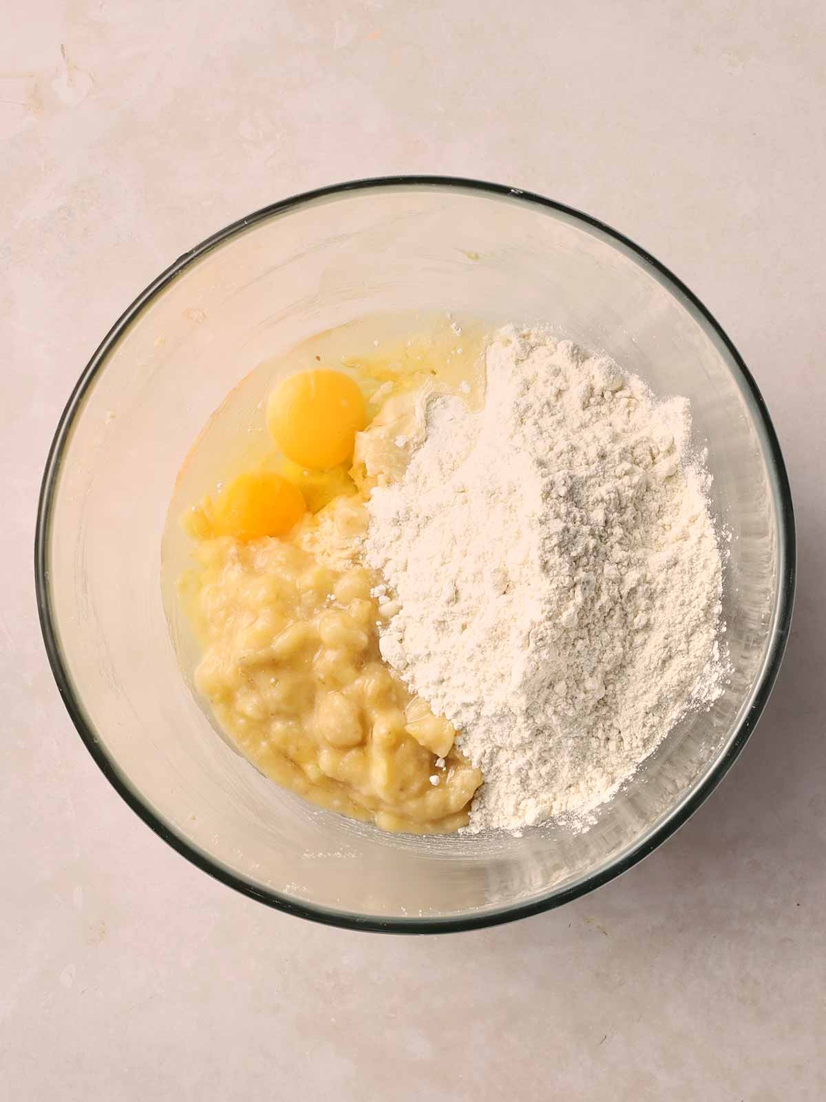 A glass bowl with cracked eggs inside, with flour and mashed banana. On a white surface.