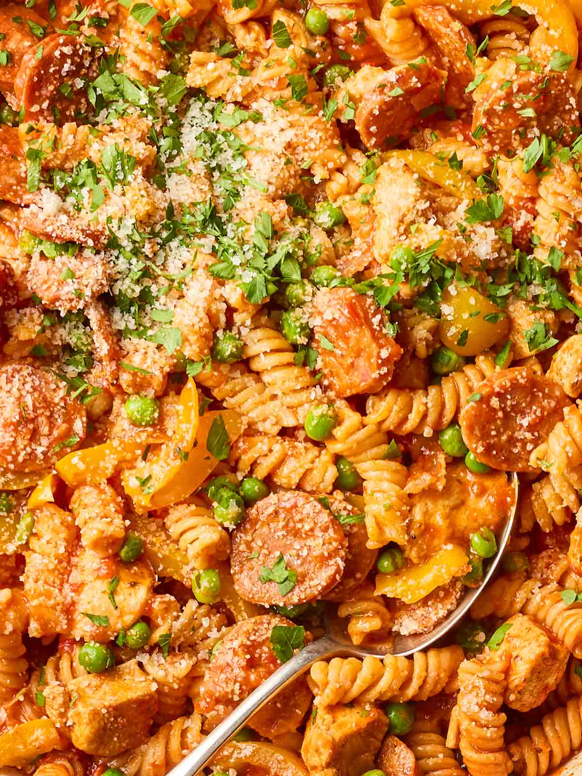 A full close up of pasta in a sauce with chicken and chorizo, with peas and parsley and parmesan, with a spoon in.