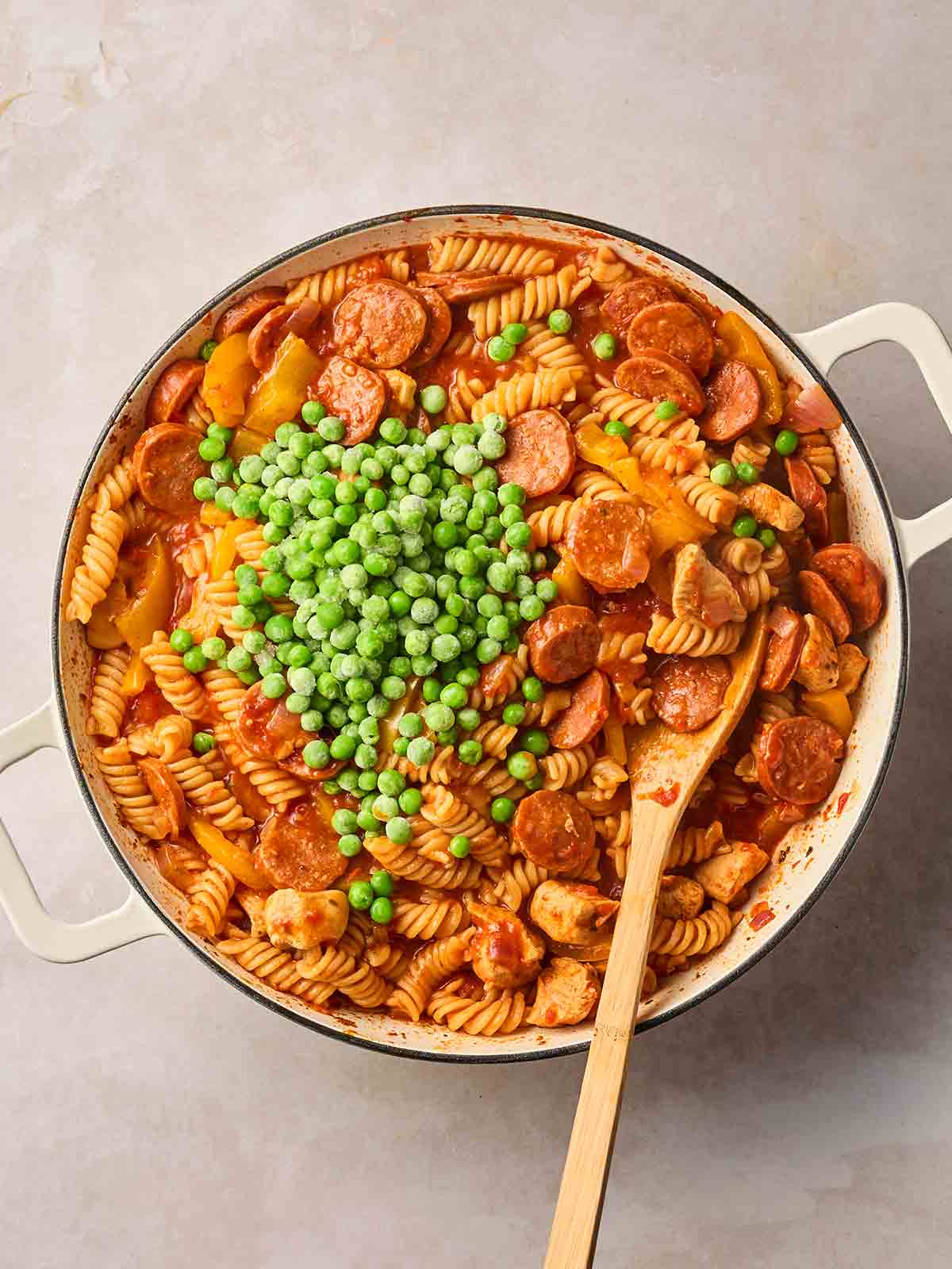 A white pan filled with chicken and chorizo pasta, with frozen peas about to be stirred in, with a wooden spoon.