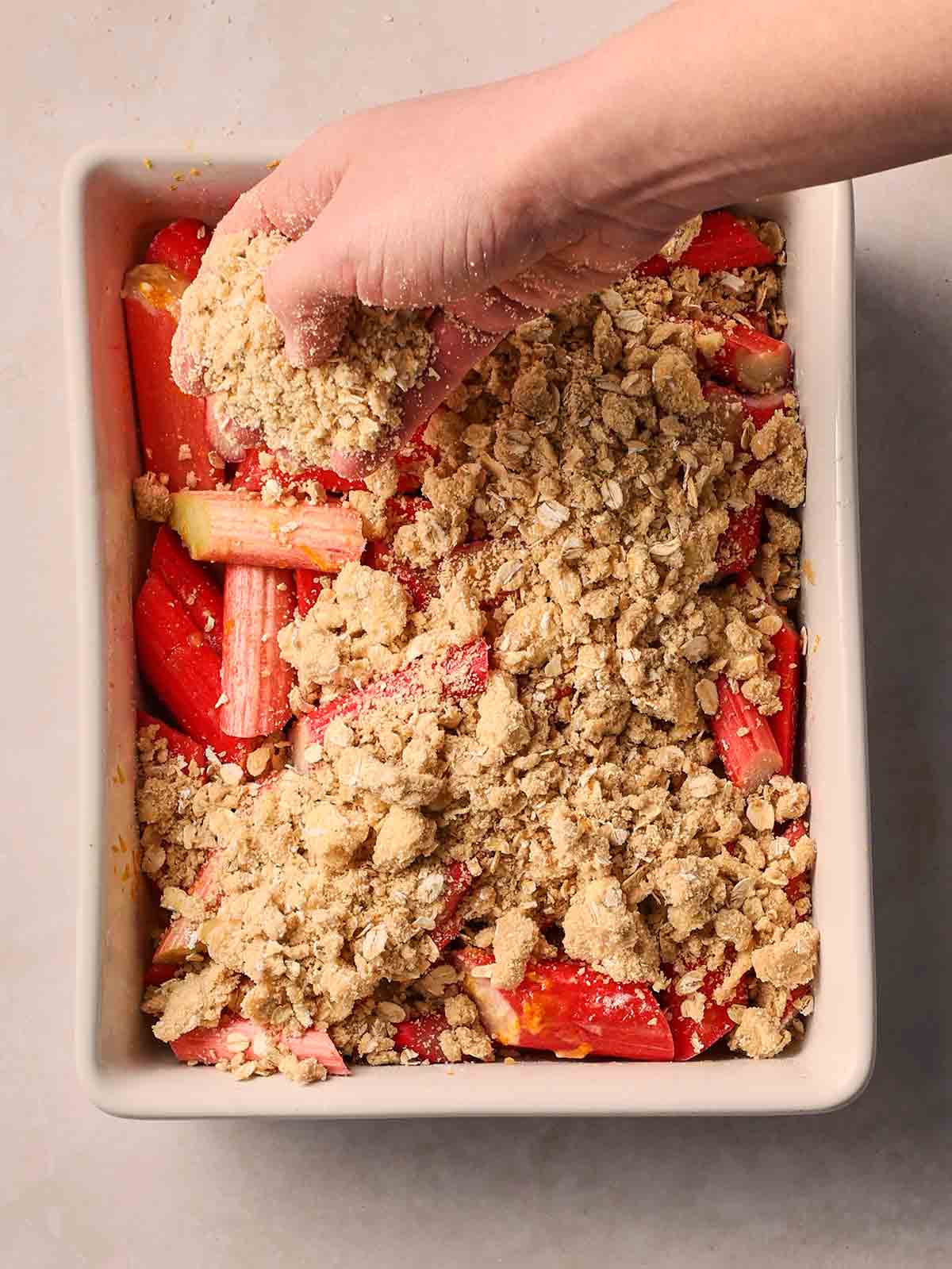 A hand sprinkling over a crumble topping of the rhubarb filling in an oven dish on a white surface.