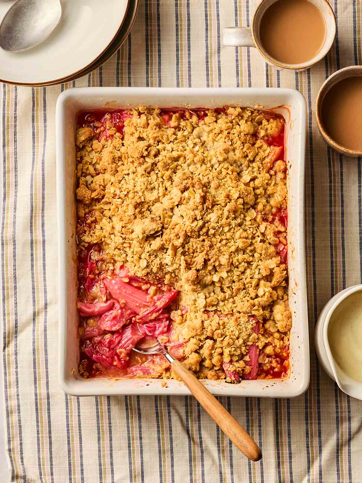A baked Rhubarb Crumble on a stripy tablecloth with cups of tea to these side and a spoon in to serve.