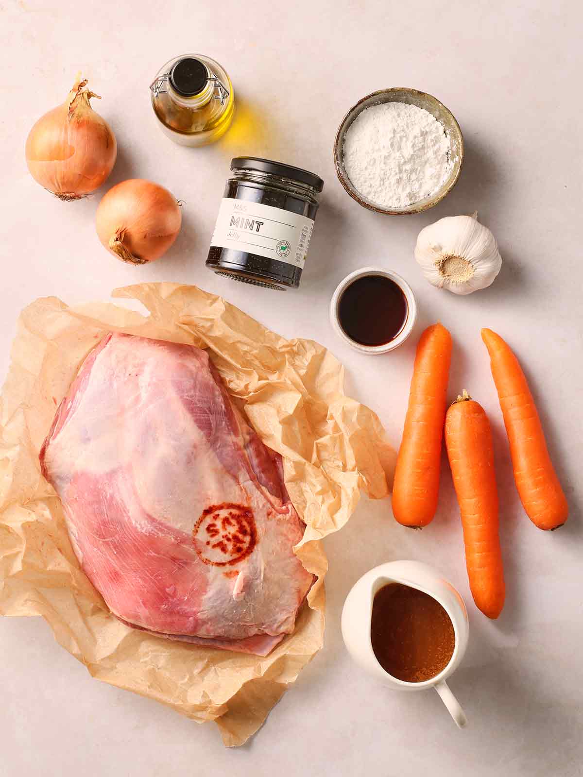 A raw lamb shoulder on a white surface with other ingredients, including carrots, stock, mint sauce, flour, onions and garlic.