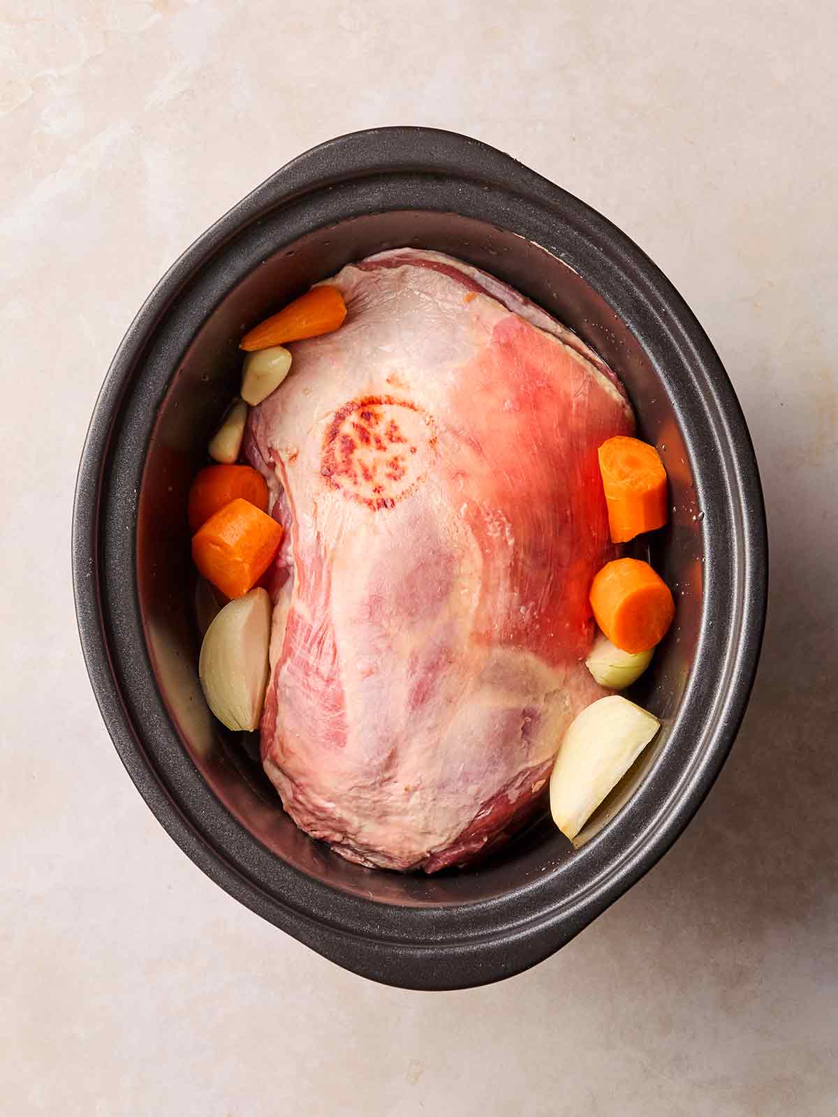 A slow cooker pan on a white surface, filled with a raw lamb shoulder joint, carrots and onions.