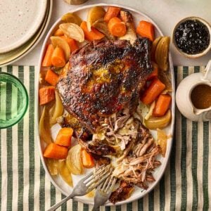 A cooked lamb shoulder joint on a platter with onions and carrots, on a set dining table, ready to be dished up.