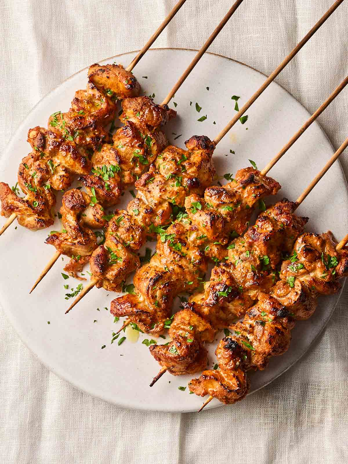 A line of skewered chicken thighs on a white plate, making charred and cooked chicken kebabs that are ready to eat.