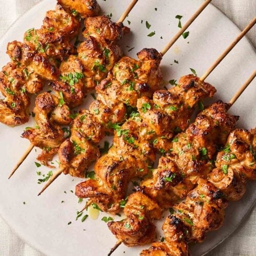 A plate with a line of cooked Chicken Kebabs on, still on the skewers, with a white linen background.