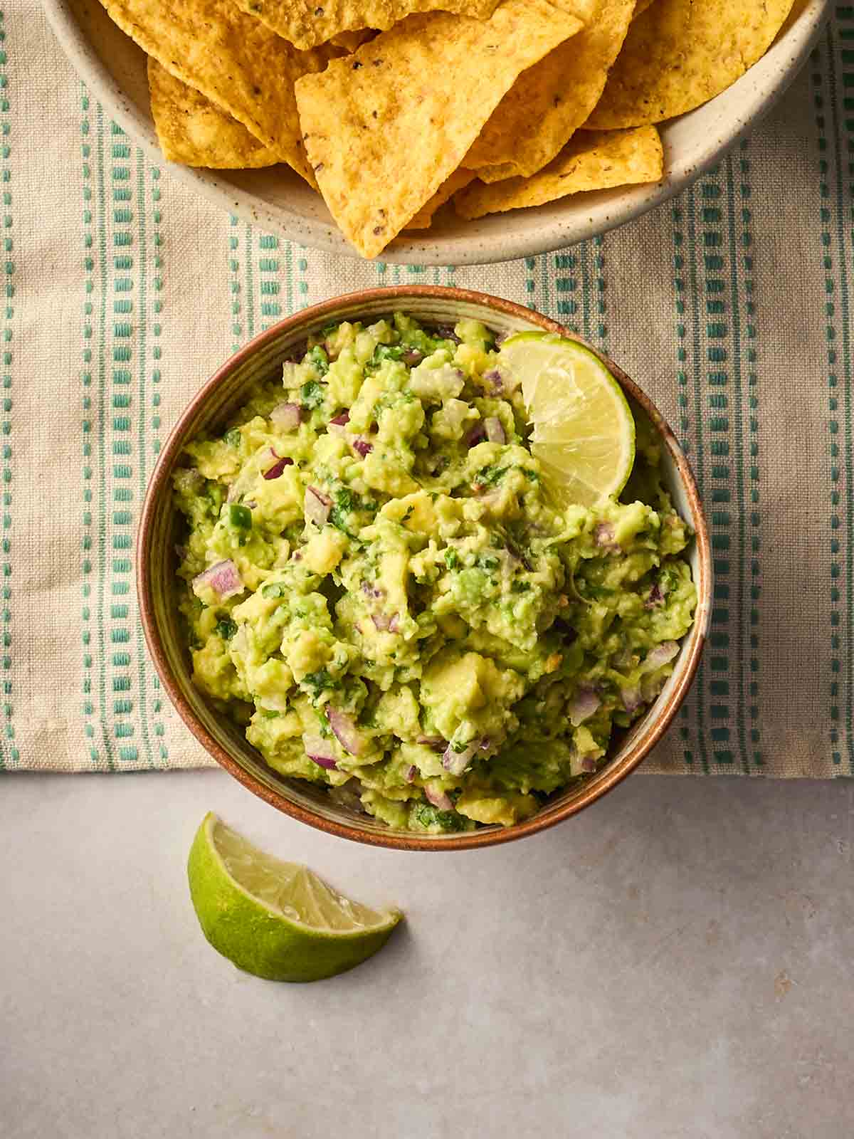 A bowl of homemade guacamole on a table with a tablecloth, and tortilla chips to the side and a wedge of lime.