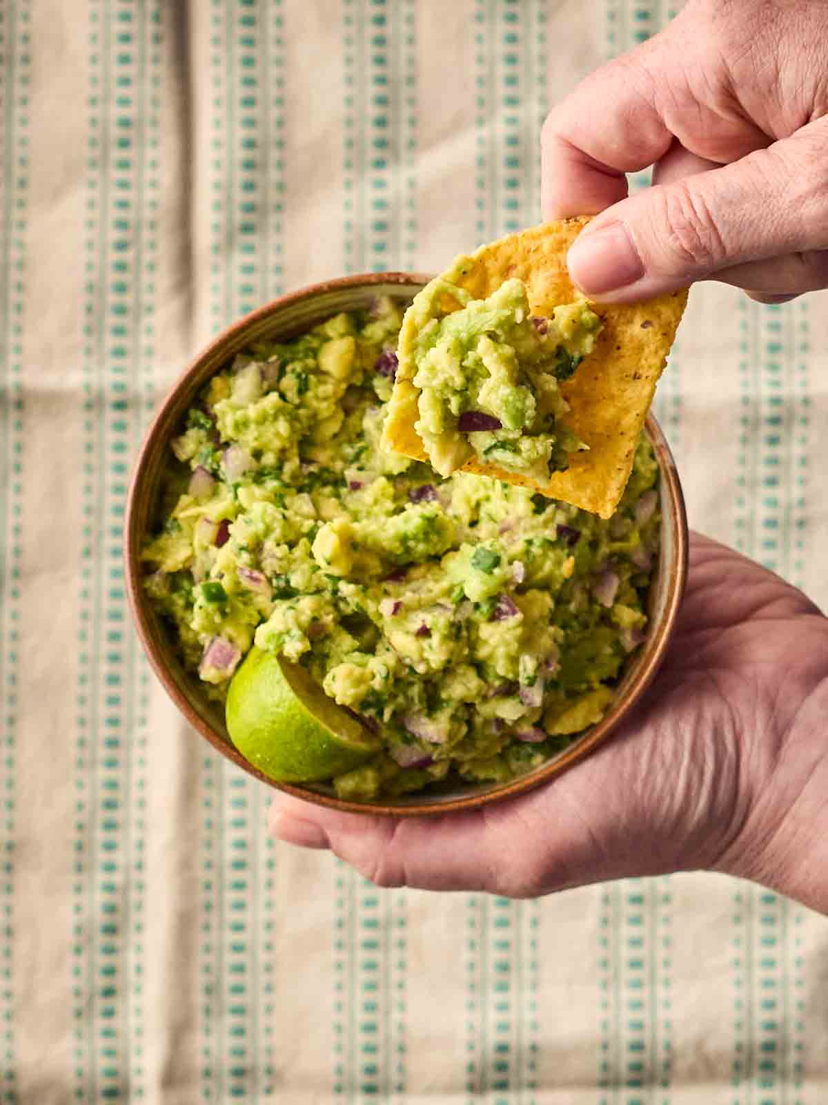 A hand holding a bowl of guacamole with another hand dipping in a tortilla chip.