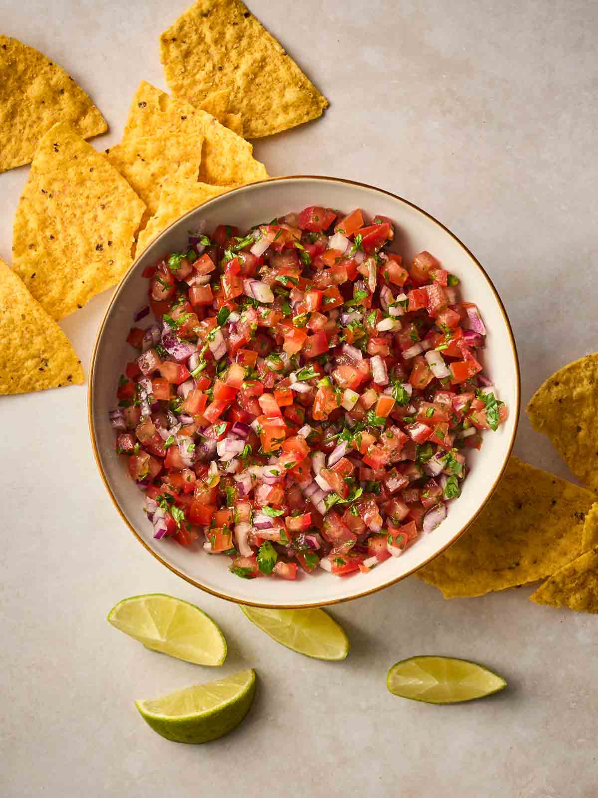 A white bowl of homemade Salsa on a white surface, with tortilla chips and lime wedges around the side.