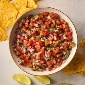 A bowl filled with fresh Salsa, with tortilla chips and lime wedges on the surface to the side.