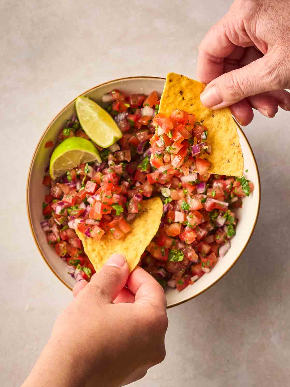 Two hands holding tortilla chips, dipping into a bowl of fresh salsa.