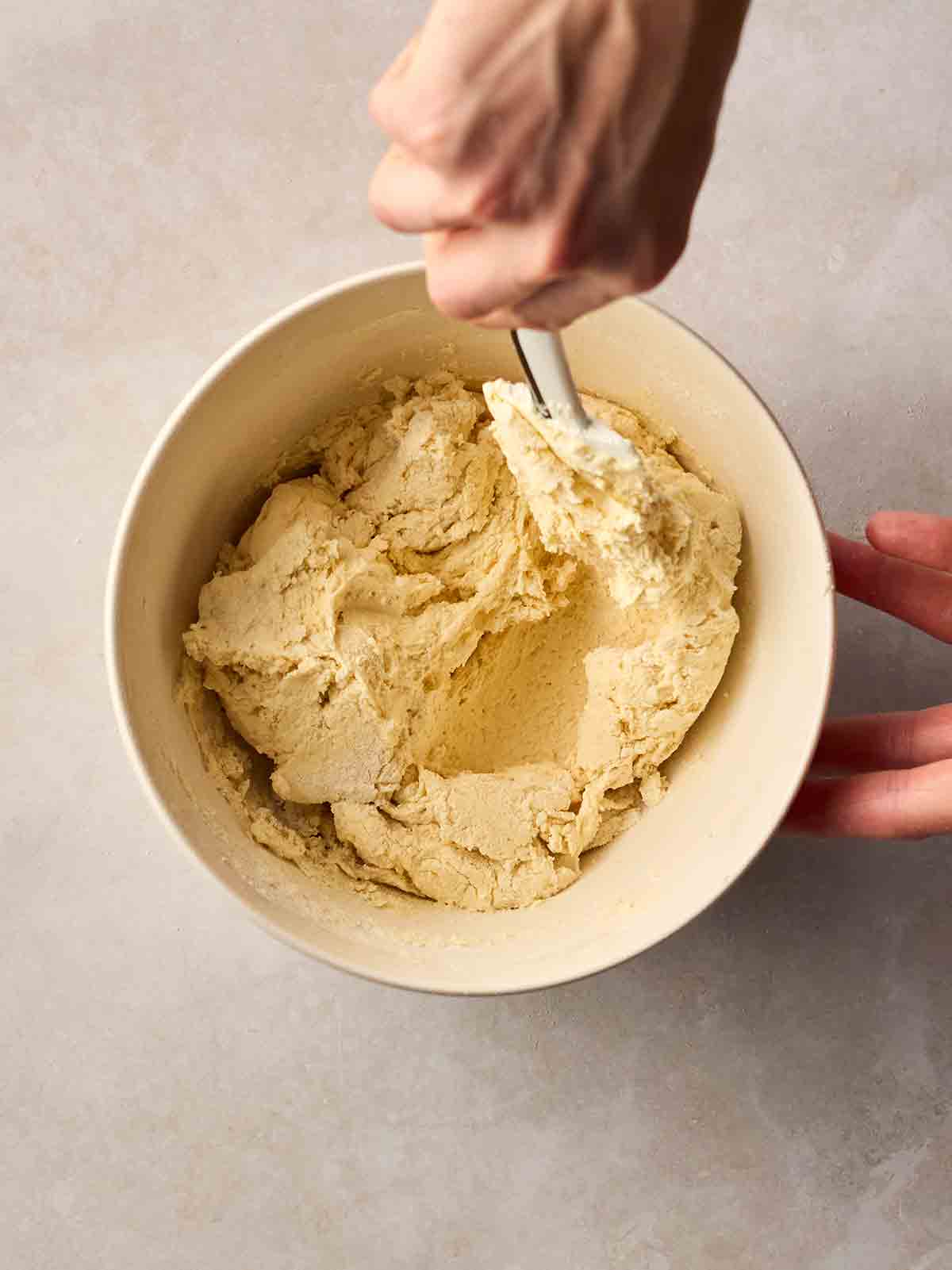 A hand mixing a cake batter with a spatula in a bowl on a white surface.
