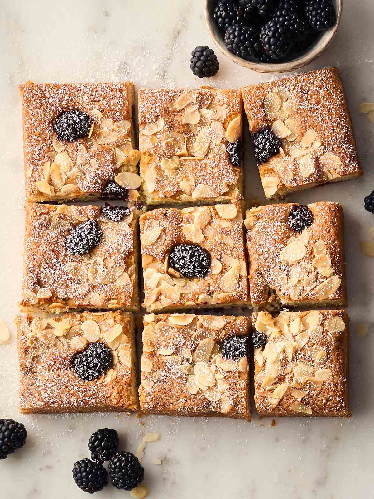 A square baked Blackberry Cake on a white surface, cut into nine squares, with blackberry, flaked almonds and a sprinkling of icing sugar on top.