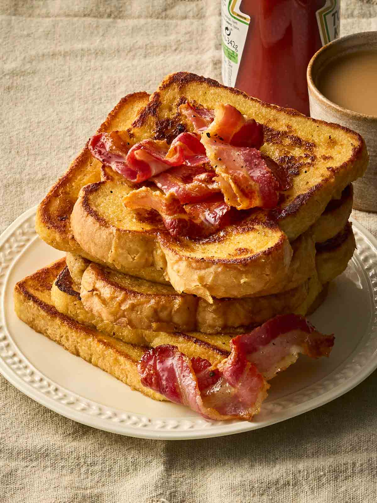 A side angle of a stack of Eggy Bread with bacon on a plate, with a cup of tea in the background.