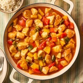 A large pot of Sweet and Sour Chicken on a green stripy tablecloth, with rice to the side, and plates and cutlery to the other side.